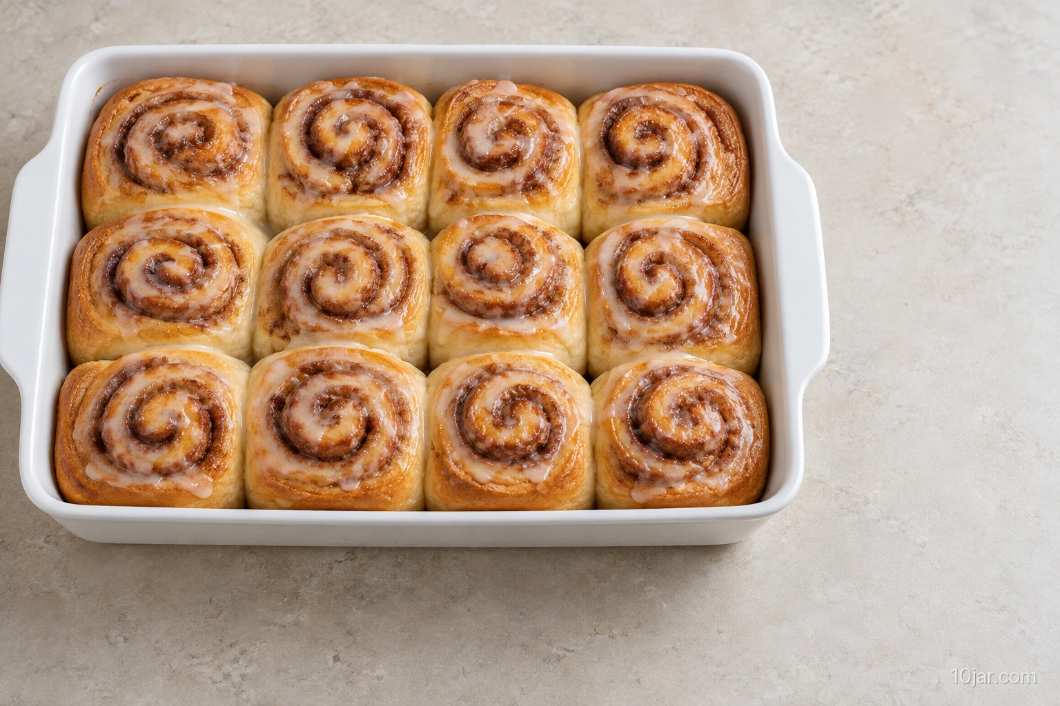 Twelve baked cinnamon rolls in a three-by-four arrangement in a white rectangular pan with a thin simple glaze